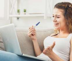 Smiling woman holding a pregnancy test and browsing on a laptop to buy HCG injections online from ReliableRx Pharmacy
