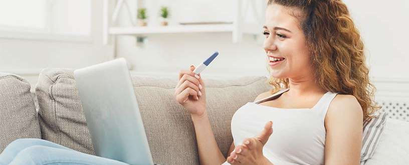 Smiling woman holding a pregnancy test and browsing on a laptop to buy HCG injections online from ReliableRx Pharmacy