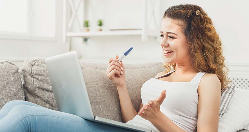 Smiling woman holding a pregnancy test and browsing on a laptop to buy HCG injections online from ReliableRx Pharmacy